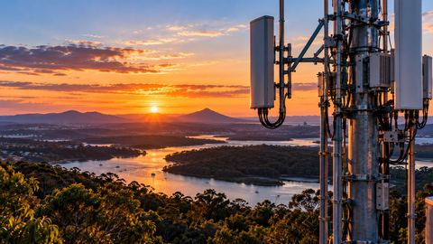 A mobile network tower above bushland and a river in Queensland, with the sun setting behind the ranges